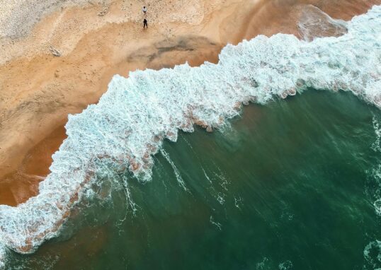 Aerial shot of ocean waves crashing onto a sandy beach with two people in the distance.